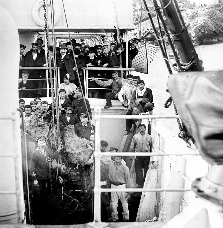 Black and white photograph: men in caps and suits crowd the deck and rails of a steamship; in the foreground a large find, crusted from the sea, hangs in a rope net